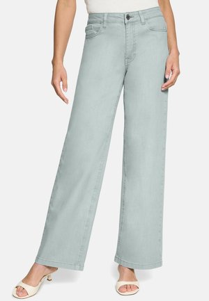 Femme portant un jean large bleu clair et des talons ouverts blancs, debout les mains détendues le long du corps.