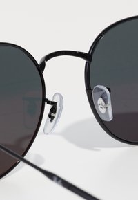 Ochelari de soare rotunzi, negri, cu lentile închise la culoare, având tampoane nazale transparente atașate la un cadru subțire din metal. Include un brat drept negru.