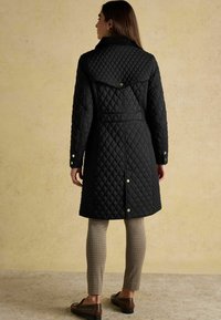 Sieviete, tērpta melnā quiltētā garajā mētelī ar spiedpogām, bēšās rūtainās biksēs un brūnos loafers, stāv uz vienkārša bēša fona.