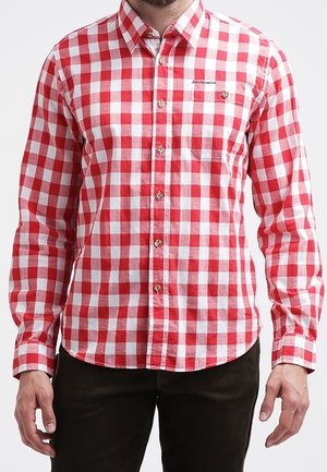 Homme portant une chemise à carreaux rouges et blancs à manches longues avec un pantalon noir, debout devant un fond uni.