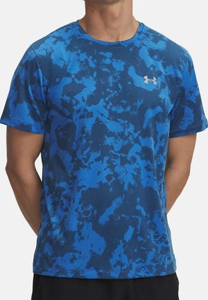Mann trägt ein kurzärmeliges, blaues Tarnmuster-Unter Armour-Sportshirt und steht mit den Händen hinter dem Rücken.