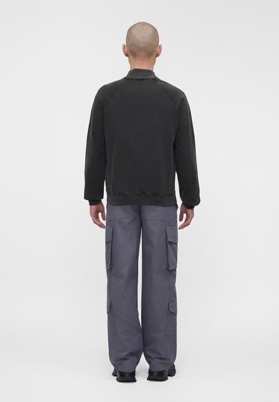 Άτομο με ξυρισμένο κεφάλι που φοράει μαύρο μακρυμάνικο πουκάμισο και άνετο γκρι cargo παντελόνι, στέκεται με την πλάτη γυρισμένη σε μια απλή λευκή επιφάνεια.