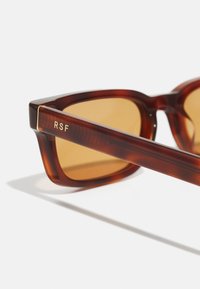 Gafas de sol de tortuga marrón con el logotipo dorado de RSF en las patillas, que cuentan con lentes tintados marrones y un diseño de marco angular. Acabado mate.