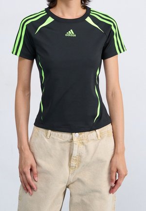Personne portant un t-shirt noir moulant Adidas avec des rayures vert néon et un pantalon beige taille haute, debout devant un fond uni.