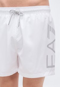 Λευκά μαγιό σορτς από ελαφρύ υλικό, με γκρι κορδόνι και διακοσμητικό λογότυπο με κουκκίδες στο πλάι. Ομαλή υφή, μήκος μέχρι το γόνατο.