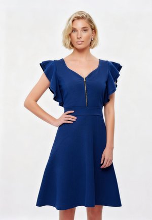 Blondynka stojąca z ręką na biodrze, ubrana w granatową, bezrękawową sukienkę z falbankami na ramionach i ozdobnym zamkiem błyskawicznym z przodu.