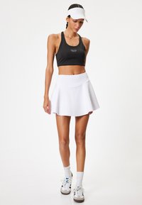 Svart sportbh med texten "FEELING GOOD", matchad med en vit utställd kjol. Modellen fulländar looken med vita strumpor och sneakers.