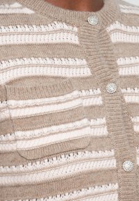 Beige Strickjacke mit horizontalen Streifen in Creme. Sie hat einen runden Ausschnitt, ein strukturiertes Material, eine Vordertasche und glänzende Knopf-Akzente.