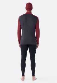 Man die blootsvoets staat met de rug naar ons toe, gekleed in een bordeauxrode en donkergrijze lange mouw top, een bordeauxrode beanie en zwarte strakke leggings op een lichte achtergrond.