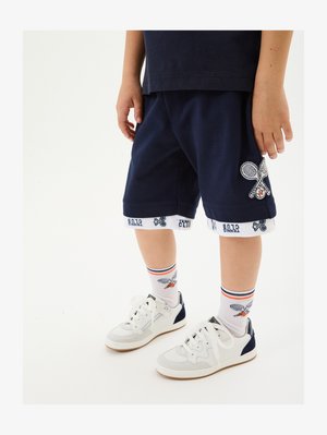 Jūras zila šorti ar tenisa raketes izšuvumu, baltā josta ar tekstu "Tennis Club". Saskaņoti ar baltām kedām un rakstainām zeķēm.