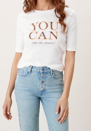 Woman wearing white shirt with "YOU CAN take the chance!" text and high-waisted light blue jeans with four visible buttons.