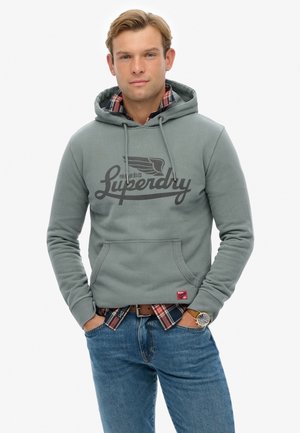 Sudadera gris con bolsillo tipo canguro y logo negro. Usada sobre una camisa de cuadros, combinada con jeans azules. El material parece suave, con puños acanalados.
