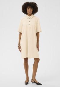 Béžové šaty s krátkym rukávom a golierom, so zapínaním na gombíky, vyrobené z hladkej tkaniny. Nosí sa s čiernymi špicatými lodičkami na podpätku.