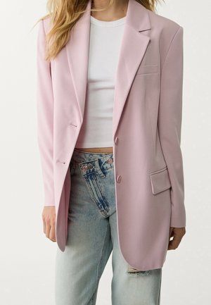 Femme portant un blazer rose clair par-dessus un top court blanc et un jean délavé taille haute avec une déchirure à l’avant.