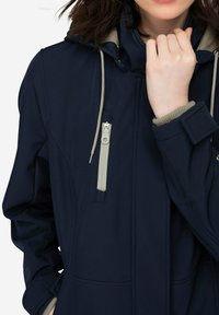 Chaqueta azul marino con capucha, con forro beige y bolsillo con cremallera. Fabricada en un tejido suave con puños elásticos y cordones ajustables.