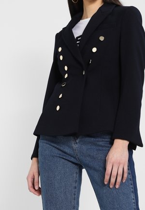 Vrouw draagt een zwarte dubbelrijige blazer met gouden knopen en blauwe jeans, staand tegen een effen lichte achtergrond.