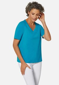 Türkise V-Neck T-Shirt aus glattem Stoff mit kurzen Ärmeln und dekorativen Nieten entlang des Ausschnitts. Kombiniert mit weißen Hosen.