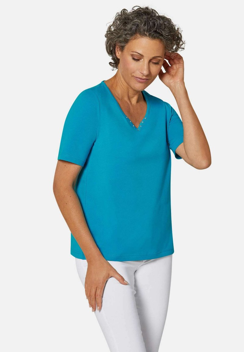 Türkise V-Neck T-Shirt aus glattem Stoff mit kurzen Ärmeln und dekorativen Nieten entlang des Ausschnitts. Kombiniert mit weißen Hosen.