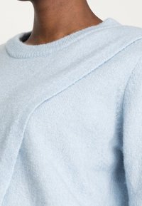Pull bleu clair en tissu doux et duveteux. Comprend un col rond côtelé et un détail de couture diagonale sur le devant.