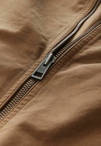 Zbliżenie na częściowo zamknięty srebrny metalowy zamek błyskawiczny na brązowej, teksturowanej tkaninie.