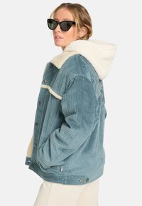 Lyseblå kordfløjljakke med en cremefarvet faux fur hætte. Funktionen inkluderer trykknapper og sidelommer, med en ribbet tekstur og broderet detalje på ryggen.
