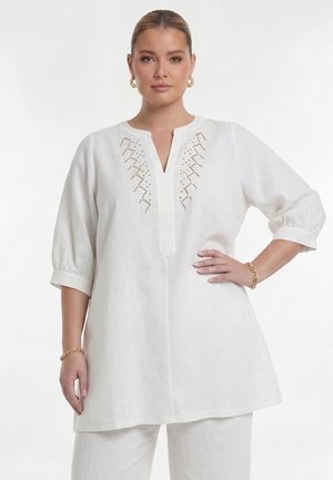 Frau trägt eine weiße Tunika mit Dreiviertelärmeln und dekorativen Perforationen am Halsausschnitt, kombiniert mit weißen Hosen und goldenem Schmuck.