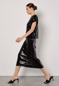 Zwarte paillettenjurk met een losse, korte mouwen top en een aansluitende rok tot aan de enkel. Draagt het met open tenen zwarte hoge hakken.