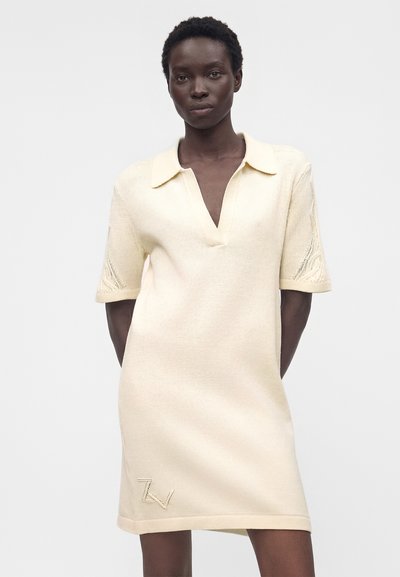 Γυναίκα που φοράει ένα κρεμ πλεκτό φόρεμα με κοντά μανίκια, γιακά και V-λαιμόκοψη, στέκεται μπροστά σε μια απλή ανοιχτόχρωμη επιφάνεια.