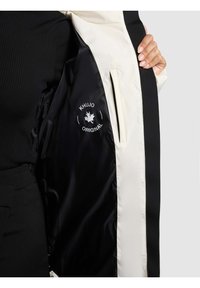 Juoda ir kreminės spalvos striukės vidus su satino tekstūra, puoštas baltu logotipo atspaudu ir šonine kišenės anga.