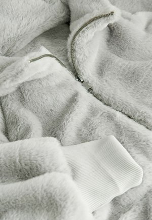 Fleece jacket in light gray with soft texture. Features a silver zipper and white ribbed cuffs. Close-up view of fabric and zipper details.