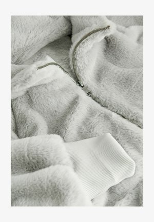 Fleece jacket in light gray with soft texture. Features a silver zipper and white ribbed cuffs. Close-up view of fabric and zipper details.