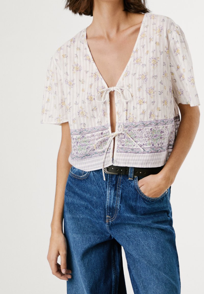 Blouse blanche courte à imprimé floral, avec un profond décolleté en V, des manches courtes, une fermeture nouée à l'avant, et une bande décorative à motifs en bas.