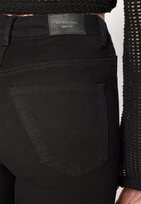 Acercamiento de unos pantalones ajustados negros con un bolsillo trasero y etiqueta de marca "perfect jeans ginatricot" combinados con una blusa de punto de malla negra.