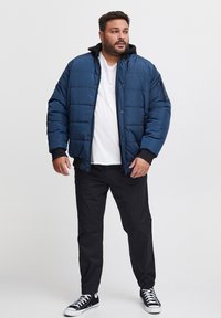 Blå polstret jakke med en sort hætte, der har en lynlåslomme, båret over en hvid T-shirt og sorte bukser, ledsaget af sorte sneakers.
