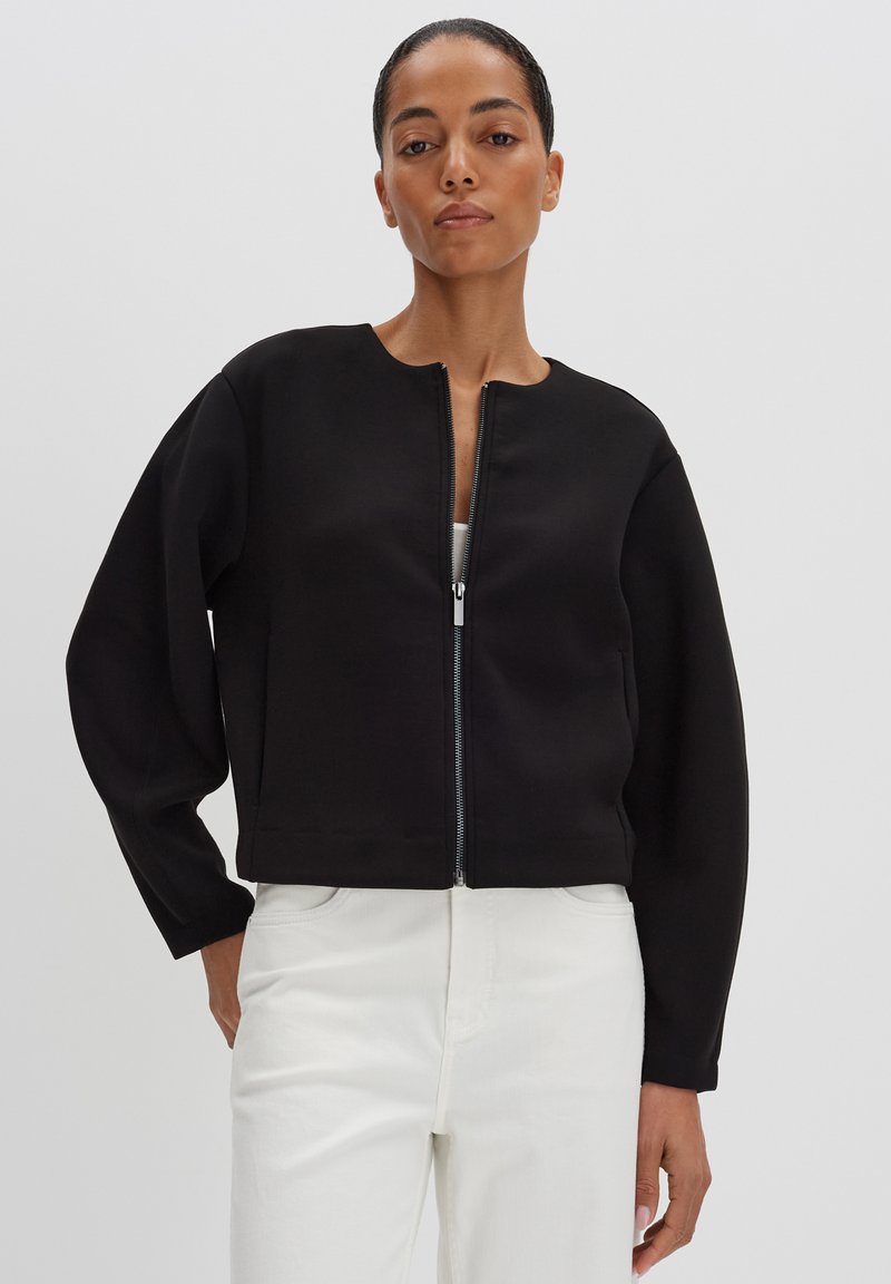 Felpa nera corta con colletto rotondo, zipper frontale e spalle scese. Tessuto morbido, abbinata a pantaloni bianchi.