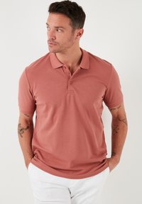 Lühikeste varrukatega roosat v ärvi polo-särk, millel on krae ja kolm nuppu. Sile tekstuur, kere järgi kohandatud disain ja õrn brändi silt äärisel.