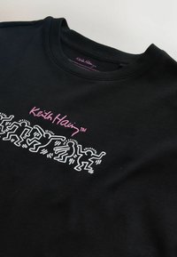 Sort T-shirt med rund hals og pink "Keith Haring" tekst samt hvide linjetegninger af dansende figurer tværs over brystet.