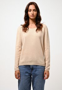 Beżowy sweter dzianinowy z okrągłym dekoltem, długimi rękawami i ściągaczem u dołu. Luźny krój z miękką teksturą, zestawiony z niebieskimi dżinsami.