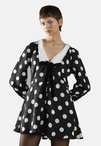 Robe noire à pois featuring un décolleté en V avec une bordure en dentelle blanche, des manches longues et un accent de nœud en velours noir à la taille.