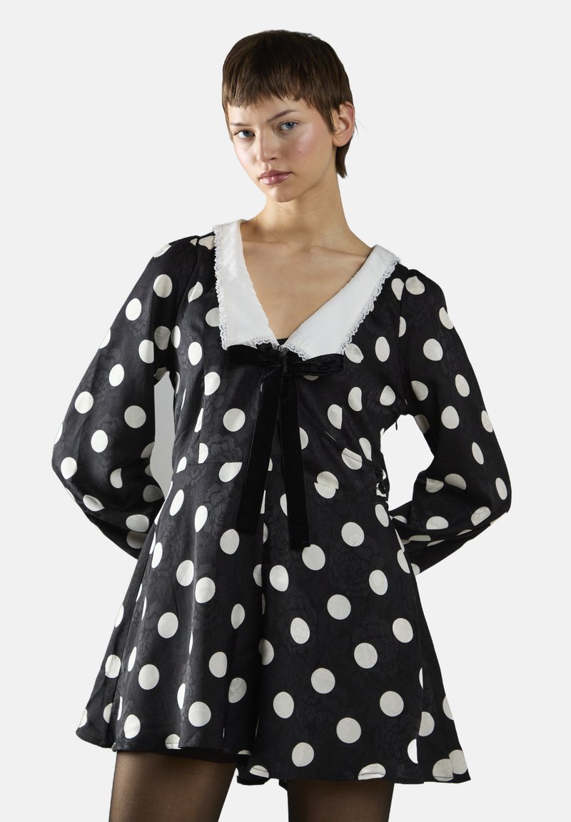 Robe noire à pois featuring un décolleté en V avec une bordure en dentelle blanche, des manches longues et un accent de nœud en velours noir à la taille.