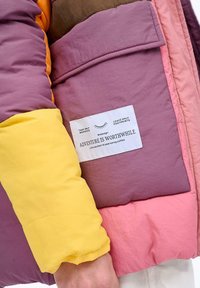 Kolorowa, wielopanelowa kurtka puchowa z sekcjami w odcieniach fioletu, różu, żółci i brązu. Etykieta zawiera tekst na białej łatce materiałowej.