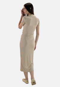 Model in een aansluitende beige jurk poseert zijwaarts, terwijl ze haar haar aanraakt, met een vervaagde achtergrond en zichtbare gouden schoenen.