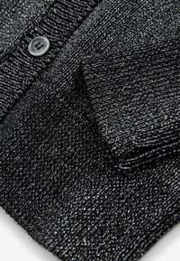 Črn teksturiran material z metalnimi nitmi, ki vsebuje podrobnosti z gumbi. Površina ima rebrast vzorec in mehak, rahlo odbleskajoč zaključek.