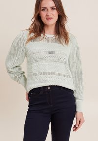 Hellgrüner gestrickter Pullover mit gemustertem Textur und langen Ärmeln, kombiniert mit dunkelblauen Jeans. Der Pullover hat eine lockere Passform und gerippte Bündchen.