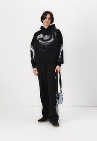 Schwarzer Kapuzenpullover mit grauem Grafikdruck und Text, kombiniert mit schwarzen Hosen. Eine kleine blaue Tasche hängt von der Schulter.
