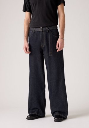 Wide-leg dark denim jeans with contrast stitching, a belt with a silver buckle, and front pockets. Paired with a black t-shirt and boots.