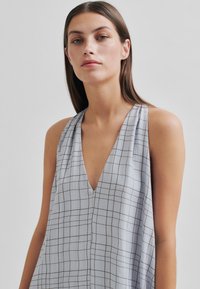 Rochie fără mâneci de culoare gri deschis, cu un decolteu adânc în V, având un model în carouri cu linii maro închis. Material ușor, croi lejer.