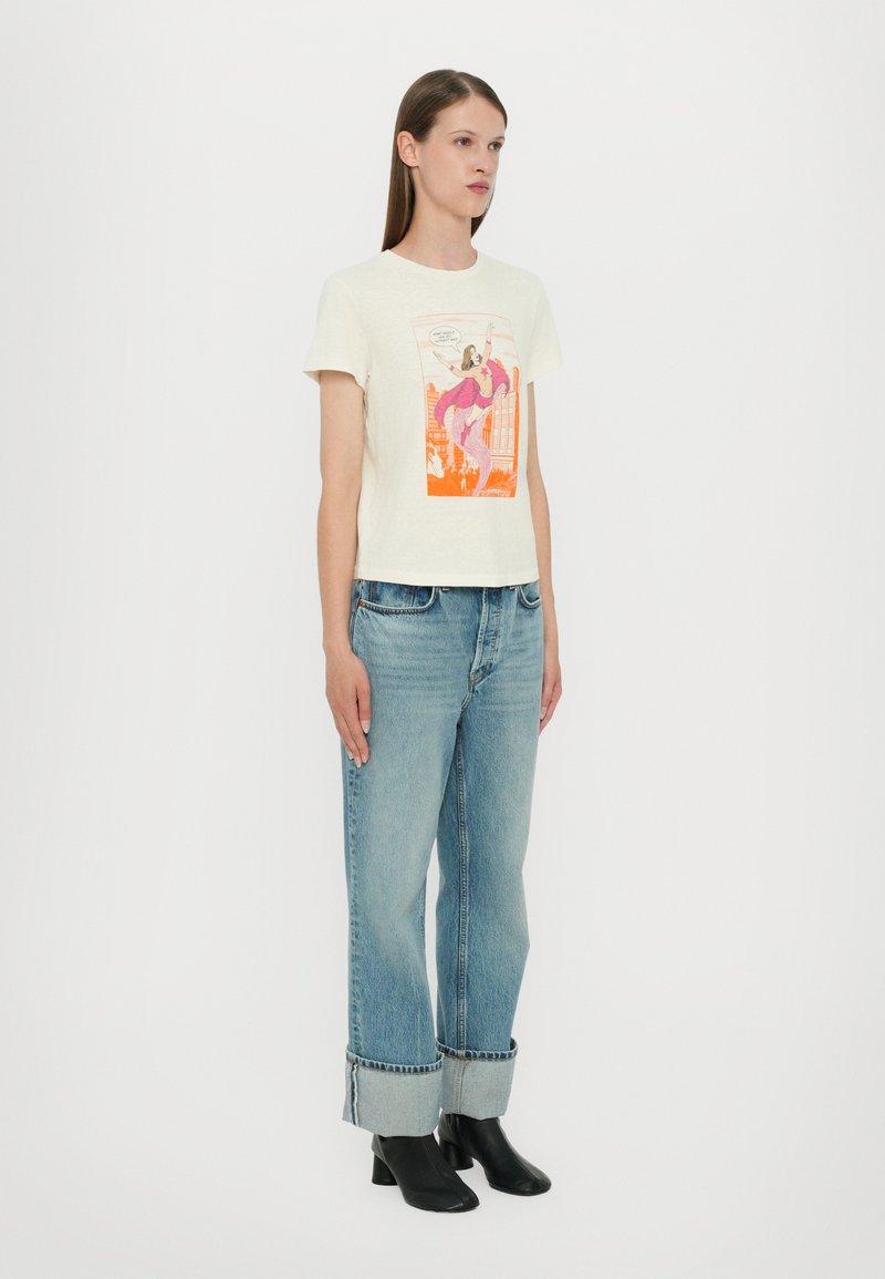 Weißes, cropped T-Shirt mit buntem Grafikdruck, kombiniert mit hellblauen Jeans mit gerader Beinführung und umgeschlagenen Säumen sowie schwarzen Stiefeletten.