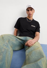 Camiseta negra de manga corta con el logo "DICKIES" en el pecho, combinada con unos vaqueros de pierna ancha en azul claro. Una gorra blanca complementa el look.