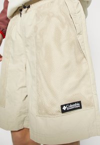 Shorts beiges Columbia Sportswear avec grande poche latérale en mesh et taille à cordon, portés avec un haut rouge à manches longues.